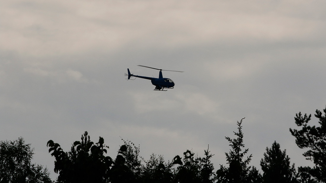 Kuvassa helikopteri ilmassa pilvistä taivasta vasten. Kuvan alareunassa näkyy puunlatvoja.