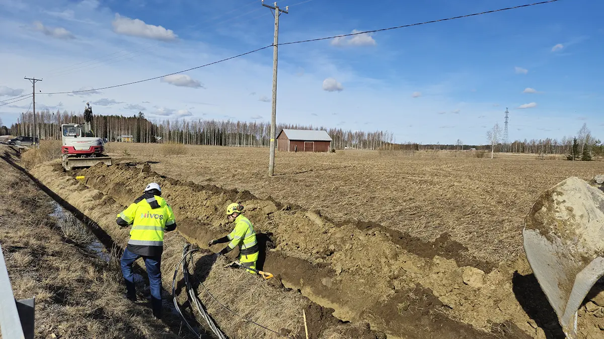 Kuva Nivoksen maakaapelointityömaalta peltomaisemassa. Taka-alalla on kaivuri työssään ja asentajat etualalla laittamassa kaapelia maahan kaivettuun uraan.