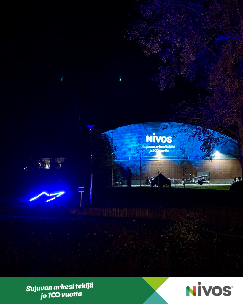 Kuva on otettu Mäntsälän jäähallin seinästä Valotapahtumassa. Seinään on heijastettu Nivoksen 100-vuotislogo valkoisella. Tausta on sininen.