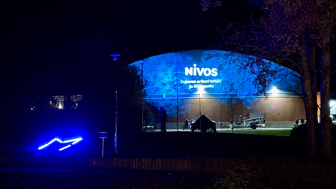 Kuva on otettu Mäntsälän jäähallin seinästä Valotapahtumassa. Seinään on heijastettu Nivoksen 100-vuotislogo valkoisella. Tausta on sininen.